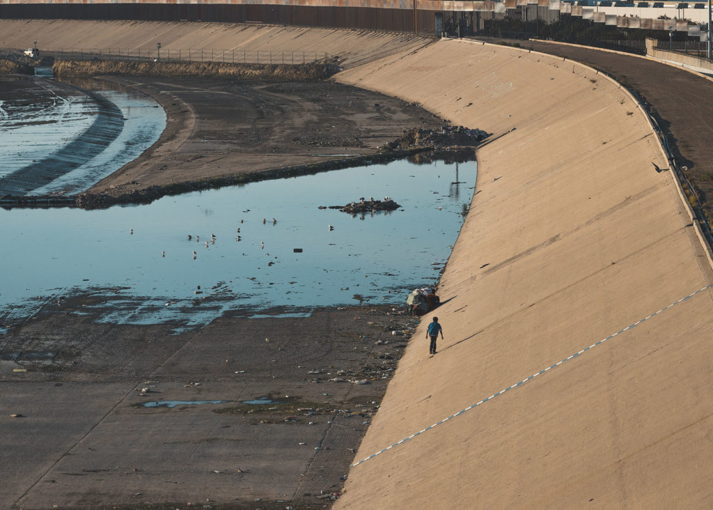 Lone South American migrant walking along the dry Tijuana waterbed, a few feet away from the United States border.